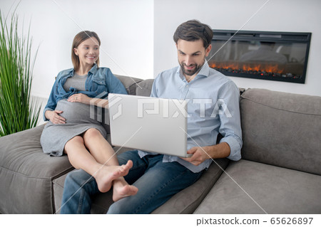 Young pregnant woman in a jeans jacket sitting near her husband while he working 65626897