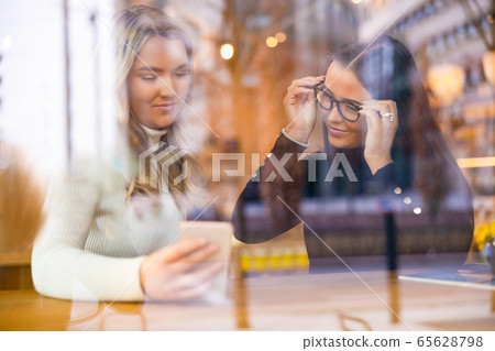 Two Beautiful Women Looking At Phone Together In Cafe Seen Through Window 65628798
