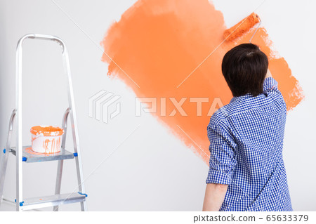 Happy middle-aged woman painting interior wall with paint roller in new house. A woman with roller applying paint on a wall. Back view. Happy middle-aged woman painting interior wall with paint roller in new house. A woman with roller applying paint on a wall. Back view. 65633379