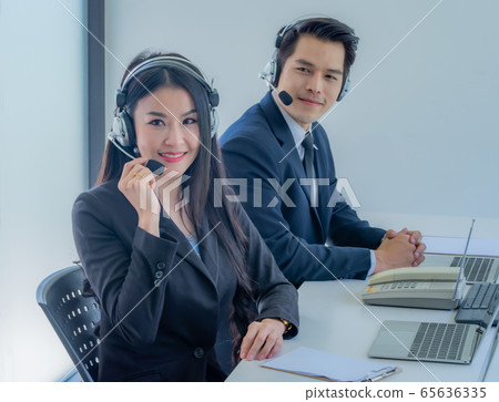 Side view of beautiful young business woman and handsome businessman in headsets using laptops while working in office. Man and woman are looking at camera. 65636335