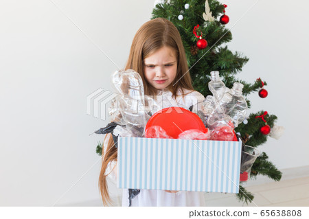 Shocked little child girl looks with opened eyes and worried expression, holding box with various plastic wastes over christmas tree background. Shocked little child girl looks with opened eyes and worried expression, holding box with various plastic wastes over christmas tree background. 65638808