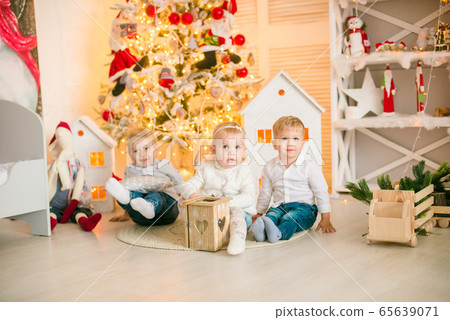 Cute little boys with blond hair plays with little girl in a bright room decorated with Christmas garlands near the Christmas tree. Happy childhood. Christmas mood Cute little boys with blond hair plays with little girl in a bright room decorated with Christmas garlands near the Christmas tree. Happy childhood. Christmas mood 65639071