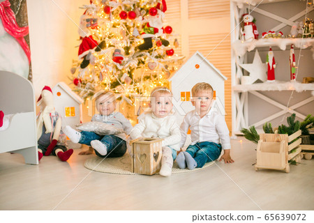 Cute little boys with blond hair plays with little girl in a bright room decorated with Christmas garlands near the Christmas tree. Happy childhood. Christmas mood Cute little boys with blond hair plays with little girl in a bright room decorated with Christmas garlands near the Christmas tree. Happy childhood. Christmas mood 65639072
