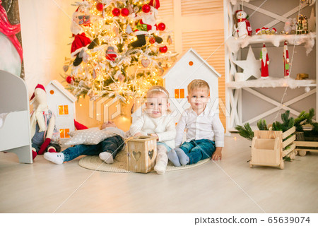 Cute little boys with blond hair plays with little girl in a bright room decorated with Christmas garlands near the Christmas tree. Happy childhood. Christmas mood Cute little boys with blond hair plays with little girl in a bright room decorated with Christmas garlands near the Christmas tree. Happy childhood. Christmas mood 65639074