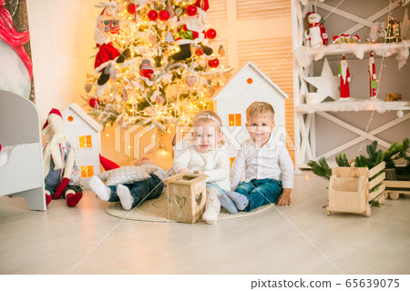 Cute little boys with blond hair plays with little girl in a bright room decorated with Christmas garlands near the Christmas tree. Happy childhood. Christmas mood Cute little boys with blond hair plays with little girl in a bright room decorated with Christmas garlands near the Christmas tree. Happy childhood. Christmas mood 65639075