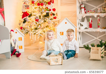 Cute little boy with blond hair plays with little girl in a bright room decorated with Christmas garlands near the Christmas tree. Happy childhood. Christmas mood Cute little boy with blond hair plays with little girl in a bright room decorated with Christmas garlands near the Christmas tree. Happy childhood. Christmas mood 65639077
