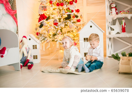 Cute little boy with blond hair plays with little girl  in a bright room decorated with Christmas garlands near the Christmas tree. Happy childhood. Christmas mood 65639078