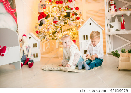 Cute little boy with blond hair plays with little girl in a bright room decorated with Christmas garlands near the Christmas tree. Happy childhood. Christmas mood Cute little boy with blond hair plays with little girl in a bright room decorated with Christmas garlands near the Christmas tree. Happy childhood. Christmas mood 65639079