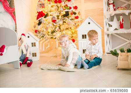 Cute little boy with blond hair plays with little girl in a bright room decorated with Christmas garlands near the Christmas tree. Happy childhood. Christmas mood Cute little boy with blond hair plays with little girl in a bright room decorated with Christmas garlands near the Christmas tree. Happy childhood. Christmas mood 65639080