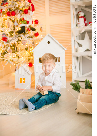 Cute little boy with blond hair plays with toys in a bright room decorated with Christmas garlands near the Christmas tree. Happy childhood. Christmas mood Cute little boy with blond hair plays with toys in a bright room decorated with Christmas garlands near the Christmas tree. Happy childhood. Christmas mood 65639083