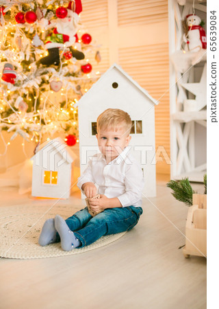 Cute little boy with blond hair plays with toys in a bright room decorated with Christmas garlands near the Christmas tree. Happy childhood. Christmas mood Cute little boy with blond hair plays with toys in a bright room decorated with Christmas garlands near the Christmas tree. Happy childhood. Christmas mood 65639084