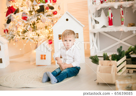 Cute little boy with blond hair plays with toys in a bright room decorated with Christmas garlands near the Christmas tree. Happy childhood. Christmas mood Cute little boy with blond hair plays with toys in a bright room decorated with Christmas garlands near the Christmas tree. Happy childhood. Christmas mood 65639085