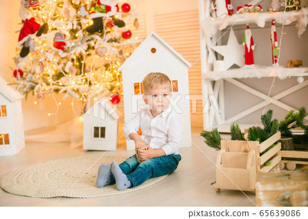Cute little boy with blond hair plays with toys in a bright room decorated with Christmas garlands near the Christmas tree. Happy childhood. Christmas mood Cute little boy with blond hair plays with toys in a bright room decorated with Christmas garlands near the Christmas tree. Happy childhood. Christmas mood 65639086