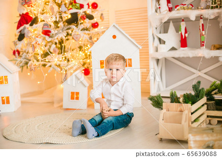 Cute little boy with blond hair plays with toys in a bright room decorated with Christmas garlands near the Christmas tree. Happy childhood. Christmas mood Cute little boy with blond hair plays with toys in a bright room decorated with Christmas garlands near the Christmas tree. Happy childhood. Christmas mood 65639088