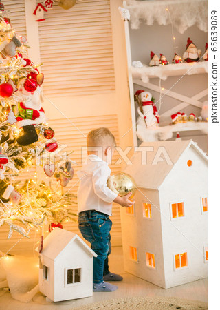 Cute little boy with blond hair plays with toys in a bright room decorated with Christmas garlands near the Christmas tree. Happy childhood. Christmas mood Cute little boy with blond hair plays with toys in a bright room decorated with Christmas garlands near the Christmas tree. Happy childhood. Christmas mood 65639089