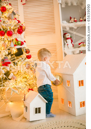Cute little boy with blond hair plays with toys in a bright room decorated with Christmas garlands near the Christmas tree. Happy childhood. Christmas mood Cute little boy with blond hair plays with toys in a bright room decorated with Christmas garlands near the Christmas tree. Happy childhood. Christmas mood 65639090
