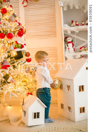 Cute little boy with blond hair plays with toys in a bright room decorated with Christmas garlands near the Christmas tree. Happy childhood. Christmas mood Cute little boy with blond hair plays with toys in a bright room decorated with Christmas garlands near the Christmas tree. Happy childhood. Christmas mood 65639091