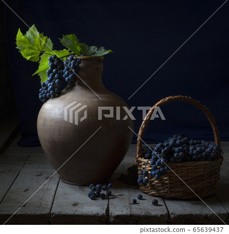 Still life with a big old jug and wild grapes. A bunch of blue grapes with bright green leaves lies in a jug. A lot of grapes lies in a wicker basket next to a jug on a wooden table. Country style. Still life with a big old jug and wild grapes. A bunch of blue grapes with bright green leaves lies in a jug. A lot of grapes lies in a wicker basket next to a jug on a wooden table. Country style. 65639437