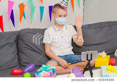 Boy in medicine face mask and festive cap celebrates birthday by video call to phone Boy in medicine face mask and festive cap celebrates birthday by video call to phone 65649366