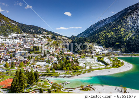 The Improbable aerial landscape of village Molveno, Italy, azure water of lake, empty beach, snow covered mountains Dolomites on background, roof top of chalet, sunny weather, a piers, coastline,  65651679