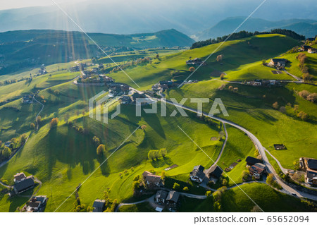 Aerial view of improbable green meadows of Italian Alps, green slopes of the mountains, Bolzano, huge clouds over a valley, roof tops of houses, Dolomites on background, sunshines through clouds 65652094
