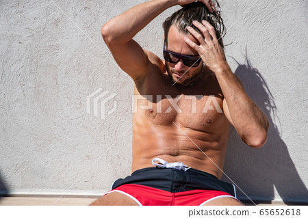 The handsome brutal man in sunglasses with a long hair and naked torso sits near the pool, collects hair in a tail, a sports suntanned body, sunglasses with a blue frame, red blue swimming shorts 65652618