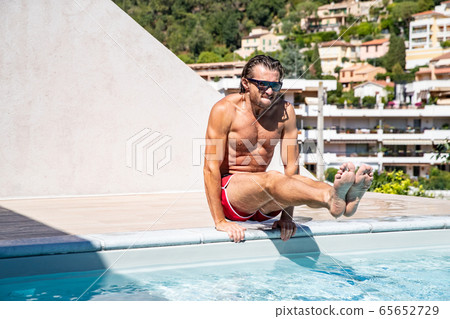 The handsome brutal man in sunglasses with a long hair and naked torso sits near the pool, Plays sports, a sports suntanned body, sunglasses with a blue frame, he is red blue swimming shorts, sunny The handsome brutal man in sunglasses with a long hair and naked torso sits near the pool, Plays sports, a sports suntanned body, sunglasses with a blue frame, he is red blue swimming shorts, sunny 65652729