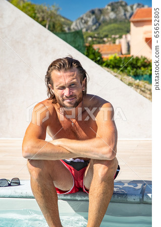 The handsome brutal man in sunglasses with a long hair and naked torso sits near the pool, is looking a camera, a sports suntanned body, sunglasses with a blue frame, he is red blue swimming shorts The handsome brutal man in sunglasses with a long hair and naked torso sits near the pool, is looking a camera, a sports suntanned body, sunglasses with a blue frame, he is red blue swimming shorts 65652805
