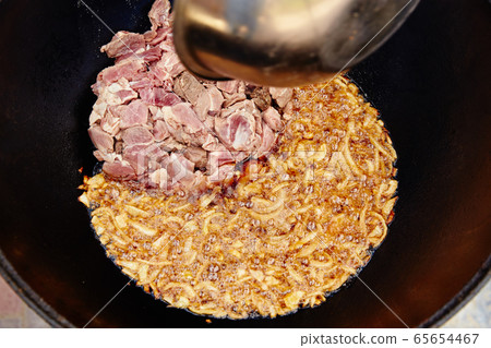 National Uzbek dish pilaf, pilaw, plov, onion and meat in big pan. Cooking process, open fire. Cooking in a cauldron on fire. Stir slowly with a skimmer. Add meat of a ram and a little he roasts 65654467