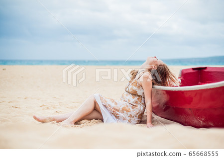 A young girl sits on the beach leaning against a boat 65668555