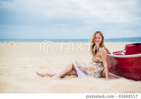 A young girl sits on the beach leaning against a boat 65668557