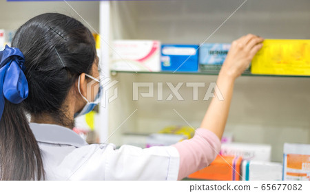 Female pharmacist taking a medicine from the shelf, wearing  lab coat during work in a modern drugstore with various pharmaceutical products.  65677082