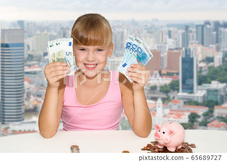 Little girl sitting on the desk with coins and pink piggy bank and holding in hands european currency  65689727
