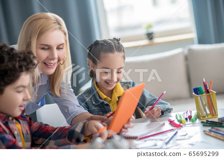 Curly boy and dark-haired girl sitting at table, blonde female bending between them, showing something on tablet 65695299