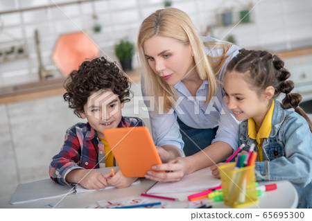 Curly boy and dark-haired girl sitting at table, blonde female bending between them, showing something on tablet Curly boy and dark-haired girl sitting at table, blonde female bending between them, showing something on tablet 65695300