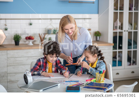 Curly boy and dark-haired girl sitting at table, doing their assignments, blonde female standing above them, helping 65695301