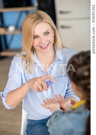 Blonde female sitting on chair, spraying antiseptic on dark-haired girl hands 65695302