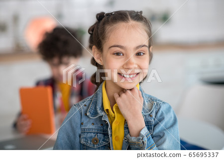 Dark-haired girl smiling into the camera, resting her chin on her hand, curly boy sitting at round table with tablet 65695337