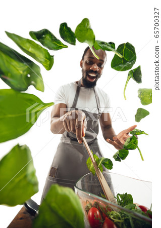 Amazing african-american man preparing unbelievable food with close up action, details and bright emotions, professional cook. Adding salad greens on the fly 65700327