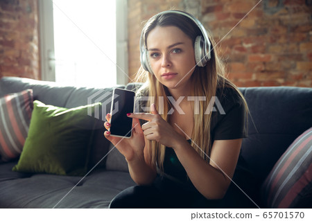Young woman wearing wireless headphones gesturing during a video conference in the living room. Pointing on phone 65701570