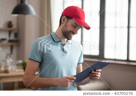 Delivery person in a red hat looking at the waybill and smiling 65703270