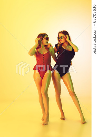 Beautiful girls in fashionable swimsuits isolated on yellow studio background in neon light. Summer, resort, fashion and weekend concept. Beautiful girls in fashionable swimsuits isolated on yellow studio background in neon light. Summer, resort, fashion and weekend concept. 65705630