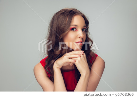 Beautiful young woman with long healthy curly hair and bright make up wearing red dress isolated on grey studio backgroud. Calm, thoughtful. 65708366