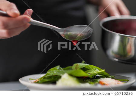 A female chef in a white uniform and a black apron in the restaurant kitchen. The cook pours red cranberry salad sauce. Making and decorating food. 65710286