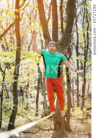 A bearded man aged walks along a slackline in the autumn forest on a sunny afternoon. The concept of Leisure and Recreation of active middle-aged people A bearded man aged walks along a slackline in the autumn forest on a sunny afternoon. The concept of Leisure and Recreation of active middle-aged people 65727299