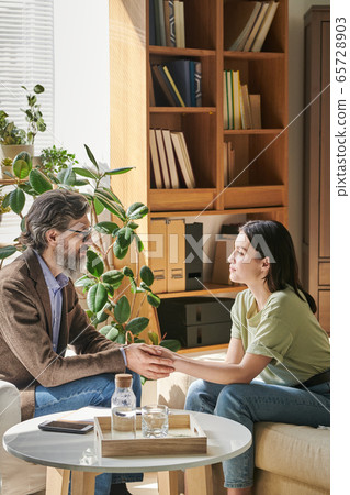 Senior professional counselor with beard holding hands of young female patient 65728903