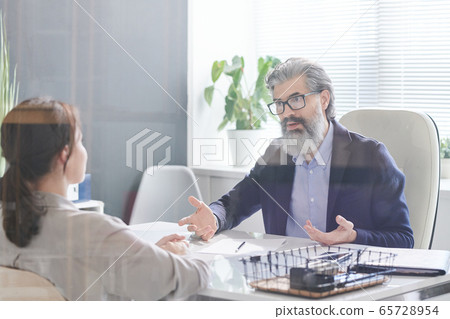 Serious employer with grey hair and beard sitting in front of applicant 65728954