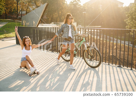 Caucasian pretty girl sits on a longboard while her girlfriend goes forward and holds her hand. 65739100