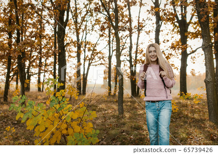Traveling girl with a backpack smiles standing on beautiful autumn forest background. Caucasian blonde travels on nature on a sunny day. Hiking concept Traveling girl with a backpack smiles standing on beautiful autumn forest background. Caucasian blonde travels on nature on a sunny day. Hiking concept 65739426