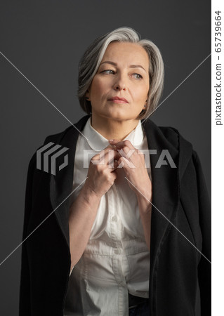 Charming gray-haired woman thoughtfully looks away adjusting collar of white shirt, throwing black jacket over her shoulders. Isolated on gray background. Lonely concept 65739664